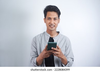 Positive Asian guy, wearing a basic shirt, using his smartphone, typing a message, browsing the internet, social media, stands on isolated white background, looking at camera, smiling
