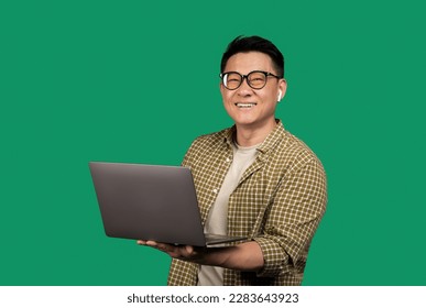 Positive asian man holding laptop computer and smiling at camera, wearing earpods and eyeglasses, posing over green background. Entrepreneurship career and internet technology concept
