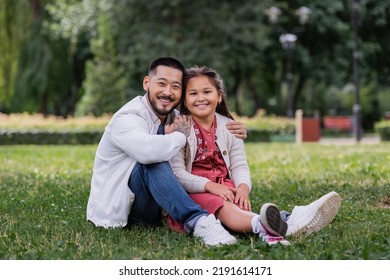 Positive Asian Man Hugging Daughter While Sitting On Grass In Summer Park