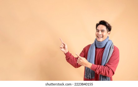 Positive Asian man in red sweater and scarf pointing fingers at ad empty space isolated on beige color background. Cheerful fashionable 20s guy aside pointing gesture - Half body length studio shot. - Shutterstock ID 2223840155
