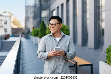 Positive asian middle aged male entrepreneur checking emails on smartphone while having break outdoors, walking by business center. Korean businessman with cellphone, copy space - Powered by Shutterstock