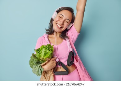 Positive Asian sportswoman dances carefree keeps arm raised up listens music via headphones dressed in spotswear carries expander embraces paper bag with vegetables moves against blue background - Shutterstock ID 2189864247