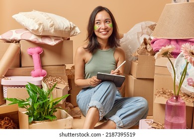 Positive Asian woman with beauty patches under eyes works on new apartment design uses modern tablet and stylus creats project poses on floor surrounded by packages with personal belongings. - Powered by Shutterstock