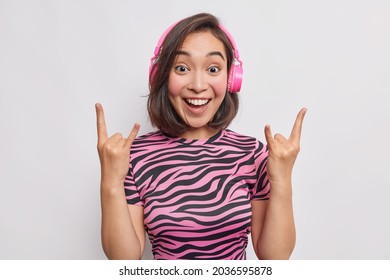 Positive Asian Woman Makes Rock Gesture Enjoys Listening Music In Wireless Headphones Shows Heavy Metal Sign Enjoys Favorite Song Has Upbeat Mood Dressed Casually Isolated Over Grey Background