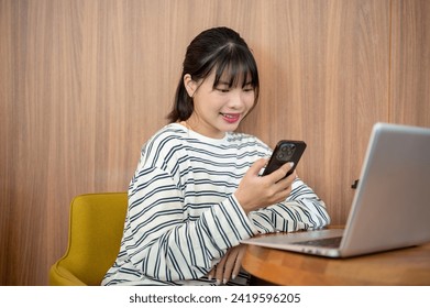 A positive Asian woman is using her smartphone while sitting at a table with her laptop in the room, reading texts or checking messages.