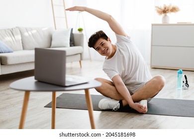 Positive athletic asian man stretching arms at home, using laptop, watching sport videos on Internet, having fitness class online with coach. Healthy lifestyle, sport on self-isolation and quarantine - Powered by Shutterstock