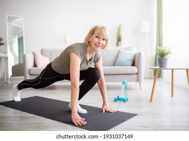 Positive Athletic Senior Woman Doing Runner's Lunge Yoga Pose On Home Workout, Copy Space. Sporty Mature Lady Stretching Her Legs, Exercising In Living Room. Domestic Training During Covid Lockdown