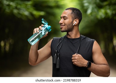 Positive athletic young black man drinking water, training at public park, using wireless earbuds, listening to music while exercising with skipping rope, copy space. Hydration and sport concept - Powered by Shutterstock