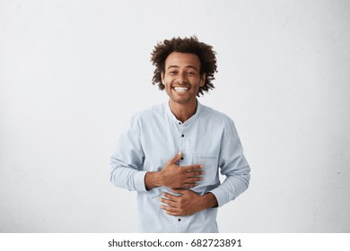 Positive Attitude And Genuine Human Emotions. Waist Up Portrait Of Cheerful Joyful Young African Man Holding Hands On Stomach While Laughing Out Loud At Something Funny, Not Suppressing His Feelings