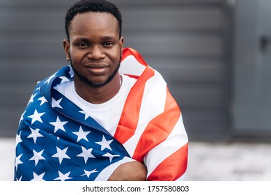 Positive attractive african american young guy wrapped himself in the flag of America and looks at the camera, outdoors - Shutterstock ID 1757580803