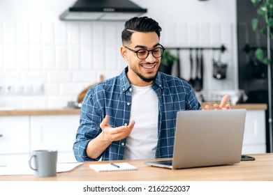 Positive Attractive Arabian Or Indian Man With Glasses, Sitting At Home In The Kitchen, Holding Video Call With Client, Virtual Meeting With Colleague, Uses Laptop, Gesturing With Hands,smile Friendly