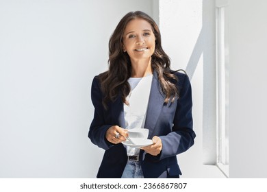 Positive attractive brunette long-haired caucasian mature lady in smart casual drinking coffee at office, stand by window with white mug in her hands, enjoy sunny day, have coffee break, copy space