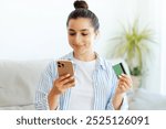 Positive attractive brunette woman, sits on a sofa in a living room, holds smartphone and credit card, enter card information for online payment in a mobile app, making online shopping, smiling