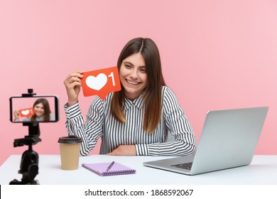 Positive Attractive Woman Blogger Holding And Showing Red Likes Counter Template, Recording Video For Subscribers On Smartphone Camera, Asking To Rate. Indoor Studio Shot Isolated On Pink Background