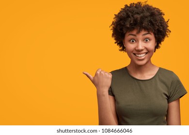 Positive attractive young female with Afro hairstyle, points aside with cheerful expression, shows something amazing at blank space, isolated over bright yellow backgroud. Advertisement and ethnicity