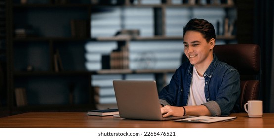 Positive attractive young guy in casual employee working on project late at night, man IT specialist sitting at desk in front of laptop, typing on keyboard, programming, panorama with copy space - Powered by Shutterstock