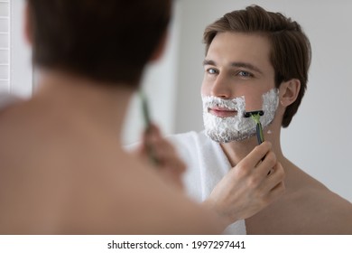 Positive attractive young guy with gel foam on lower face shaving with razor at mirror. Handsome man enjoying morning bath routine, caring for appearance, healthy facial skin. Hygiene, skincare - Powered by Shutterstock