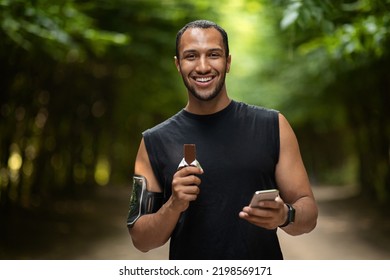 Positive attractive young middle eastern guy sportsman having break while exercising at public park, using cell phone and eating protein bar, checking workout results on smartphone, copy space - Powered by Shutterstock