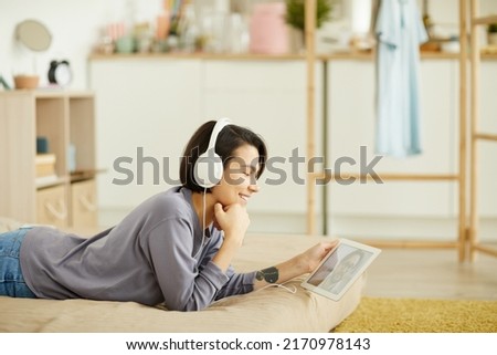 Positive attractive young woman in wired headphones lying on bed and using digital tablet while video calling boyfriend