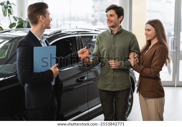 Positive auto showroom\
manager giving car key to happy young clients at car dealership.\
Millennial Caucasian couple purchasing or renting new vehicle at\
automobile store