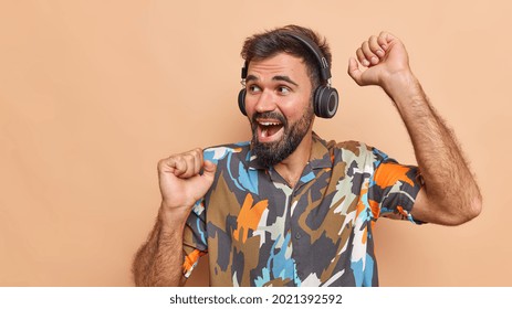 Positive Bearded Adult Man Dances Carefree Keeps Arms Raised Feels Upbeat Listens Music Via Headphones Wears Colorful Shirt Looks Away Isolated Over Brown Background Copy Space On Left Side.