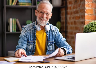 Positive Bearded Aged Man In Trendy Clothes Smiling And Looking At Camera While Writing In Notepad During Work In Home Office
