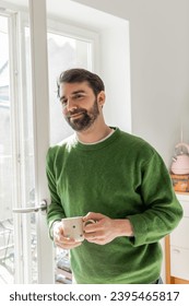 Positive bearded and brunette man in green jumper holding cup of coffee while standing near window - Shutterstock ID 2395465817