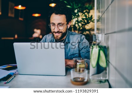 Positive bearded hipster student watching funny tutorial online in earphones connected to laptop device sitting in cafe.Young man laughing while reading news on website and enjoying music via app