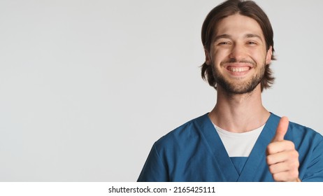 Positive bearded male doctor smiling keeping thumb up over white background. Attractive intern dressed in uniform looking confident showing approved sign isolated. Like it gesture. Copy space