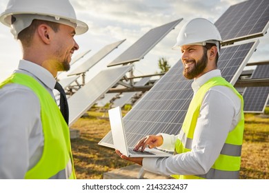 Positive bearded male engineer in hardhat and waistcoat using netbook for monitoring work of solar panels and speaking with colleague about renewable energy