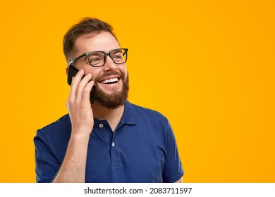 Positive bearded male hipster in glasses having phone call via cellphone on yellow background in studio and looking away
