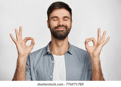 Positive bearded male model wears denim shirt, makes ok gesture and closes eyes, has happy expression, isolated over white background. Optimistic male expresses his approval. Body language concept - Shutterstock ID 776971789