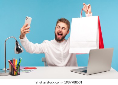 Positive bearded man blogger making selfie or recording video, posing at smartphone camera with shopping bags, bragging with purchases. Indoor studio shot isolated on blue background - Shutterstock ID 1987988531