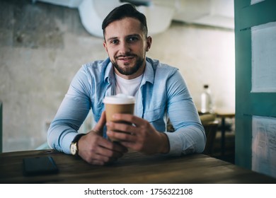 Positive bearded man in casual clothes sitting in chair at wooden table in modern cafe with cup of hot coffee while looking at camera - Shutterstock ID 1756322108
