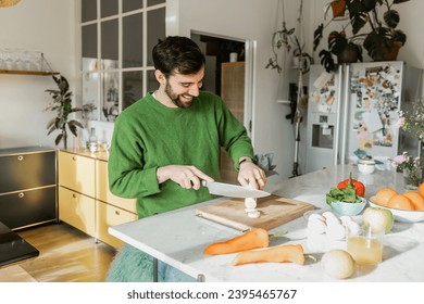 Positive bearded man in green jumper cutting fresh leek near orange juice in kitchen at home - Shutterstock ID 2395465767