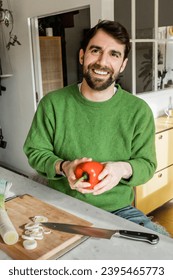 Positive bearded man in green jumper holding fresh bell pepper in kitchen at home - Shutterstock ID 2395465773