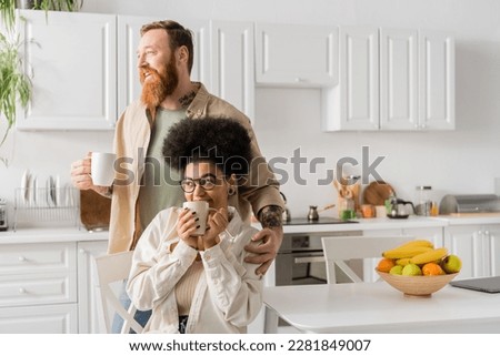 positive bearded man holding cup of coffee and hugging african american girlfriend in kitchen