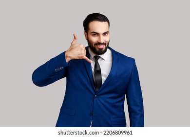 Positive Bearded Man Standing With Telephone Hand Gesture And Smiling To Camera, Flirting Offering To Contact By Phone, Wearing Official Style Suit. Indoor Studio Shot Isolated On Gray Background.