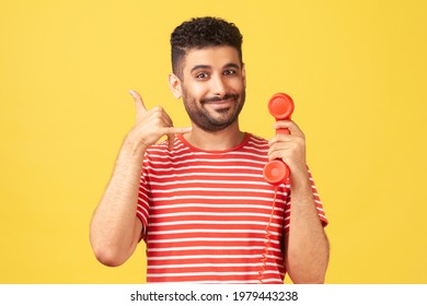 Positive Bearded Man In Striped T-shirt Holding Red Headset Of Retro Landline Telephone And Showing Call Me Gesture, 24.7 Call Centre. Indoor Studio Shot Isolated On Yellow Background