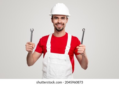 Positive bearded repairman in overall and hardhat demonstrating metal wrenches while representing professional hand tools against white background