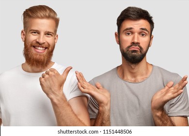 Positive Bearded Young Red Haired Male With Pleased Expression Indicates With Thumb At His Puzzled Male Friend Who Clasps Hands And Can`t Make Decision What To Do, Isolated Over White Background