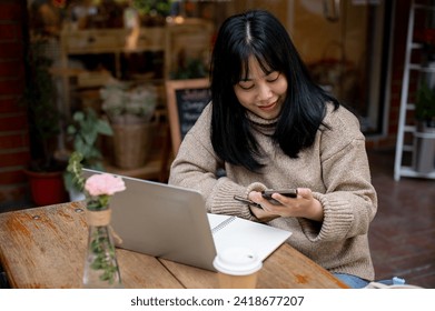 A positive, beautiful Asian woman in a cosy sweater is responding to messages on her smartphone while working remotely at a coffee shop in the city. People, wireless technology, and city life concepts