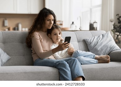 Positive beautiful brunette mom teaching sweet child girl to use online service on smartphone, relaxing on comfortable home sofa, holding mobile phone, hugging kid - Powered by Shutterstock