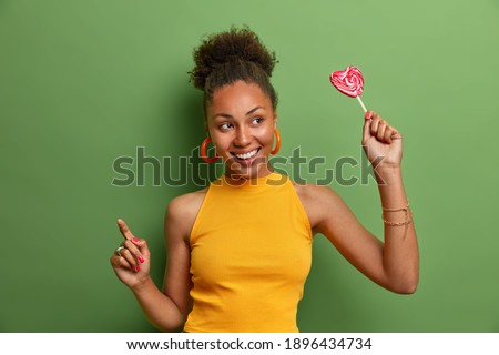 Positive beautiful curly haired woman with toothy smile holds lollipop has fun and dances carefree wears casual yellow shirt isolated over green background. Millennial girl poses with tasty candy