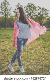 Positive Beautiful Hipster Woman In Park. Looking At Camera. Positive Human Emotion Facial Expression Body Language, Concept Of Funny Girl. Dressed In A Gray T-shirt, Blue Ripped Jeans