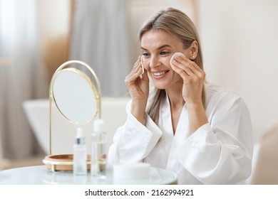 Positive Beautiful Lady In White Bathrobe Sitting In Front Of Mirror In Bathroom, Erasing Make Up From Her Face, Using Cotton Pads And Cleansing Lotion, Side View, Closeup Photo