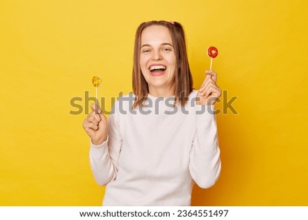 Positive beautiful woman with ponytail in jumper holding lollipop isolated over yellow background with toothy smile holds lollipop has fun and dances carefree poses with tasty candy