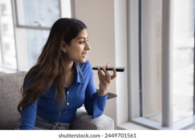 Positive beautiful young adult Indian girl recording voice message on telephone, holding smartphone at mouth, talking on speaker on cell, activating smart home application, looking away - Powered by Shutterstock