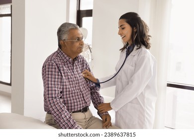 Positive beautiful young doctor examining senior Indian man, touching hand for pulse checking, applying stethoscope to chest, listening to heartbeat, smiling, asking about heart problems - Powered by Shutterstock