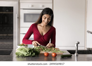 Positive beautiful young Indian chef woman preparing organic salad for dinner, caring for healthy natural nutrition, raw food diet, chopping green leaves on board, smiling, enjoying cooking - Powered by Shutterstock